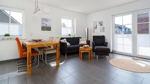 A spacious living area with a wooden dining table, a black sofa, a black armchair, and a small wooden coffee table. There are two windows providing natural light and a view of the outside. - Löwenzahn - Apartment (70sqm) for 4 persons with terrace and sep. Guest WC (Krummhörn)