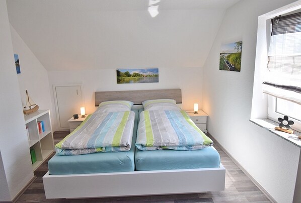 A bedroom with two beds, a nightstand, a bookshelf, and a window with blinds.