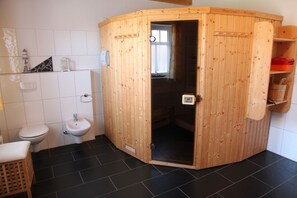 Une petite salle de bain compacte comprenant un sauna en bois, des toilettes, un lavabo et une petite étagère avec des articles de toilette.