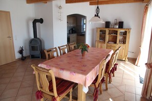 Une salle à manger avec une table recouverte d’une nappe à motifs floraux, des chaises en bois, une plante en pot et, au fond, une cuisine.