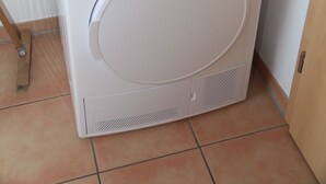 Una lavatrice bianca Sharp in un angolo di una stanza con pavimento piastrellato.