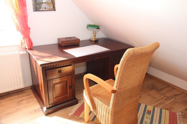 A wooden desk with a wicker chair, a radiator, a framed picture on the wall, and a lamp.