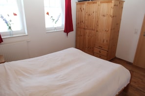 A bedroom with a bed, a wooden wardrobe, a window with a view of flowers, and a radiator.