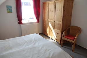 A bedroom with a bed, a wooden wardrobe, a wicker chair, and a window with red curtains.