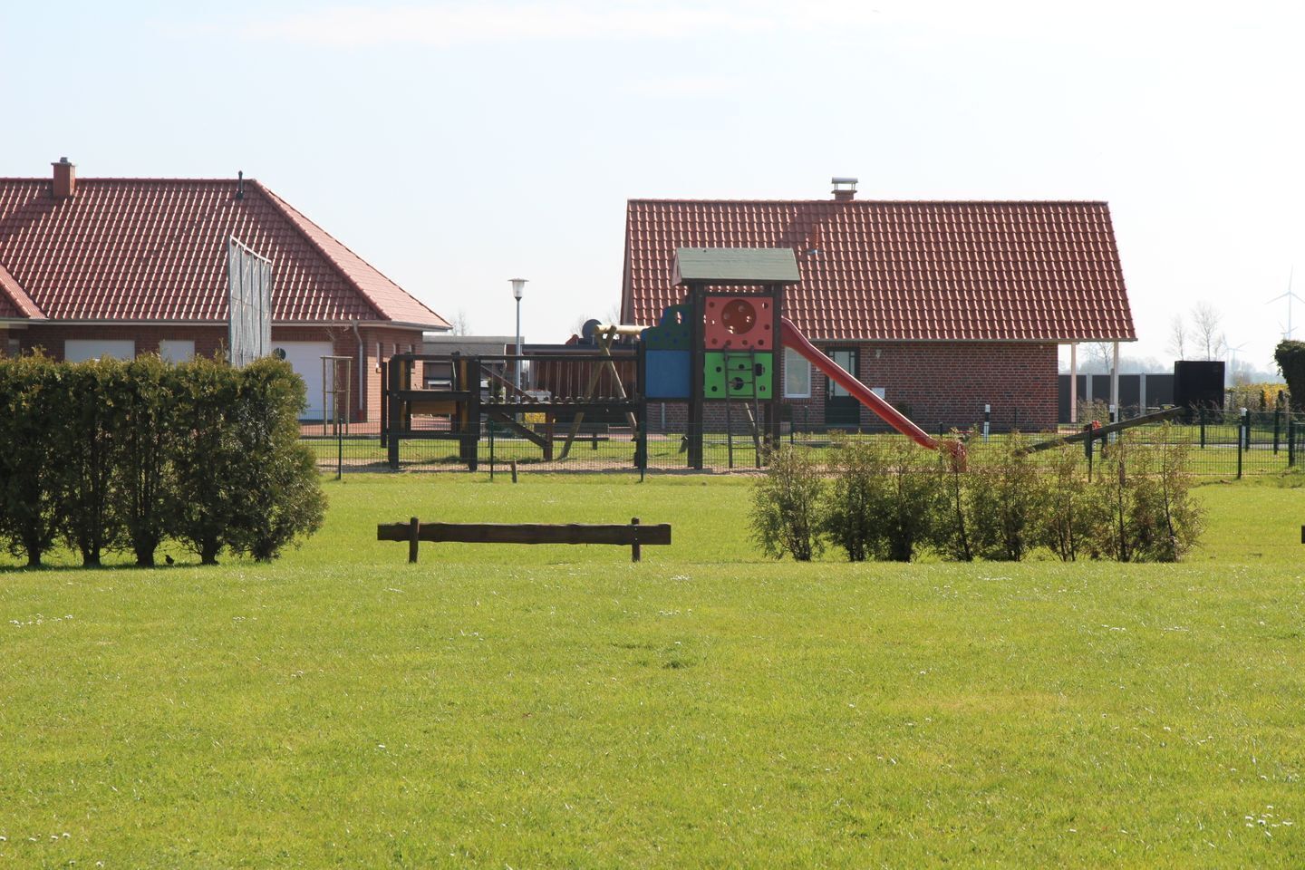 Un parque infantil con un tobogán y estructuras para escalar, rodeado de un campo de césped y un edificio con tejado de tejas rojas.