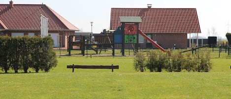 Un parque infantil con un tobogán y estructuras para escalar, rodeado de un campo de césped y un edificio con tejado de tejas rojas.