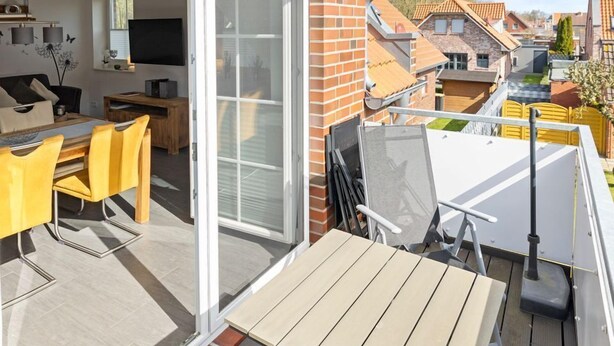 Un balcón con mesa y sillas, una puerta corredera de vidrio que da a una habitación con televisor, y vistas a casas vecinas.