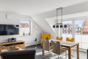 Una sala de estar moderna con un televisor de pantalla plana, muebles de madera, una mesa de comedor con sillas y ventanas amplias que ofrecen vistas del barrio.