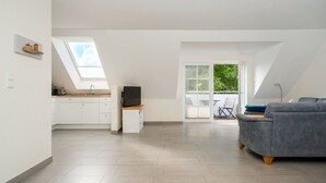 Amplia sala de estar con un sofá gris, una cocina con armarios blancos, un televisor sobre un mueble de madera y un balcón con vistas a zonas verdes.