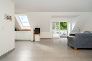 A spacious living area with a grey sofa, a kitchen with white cabinets, a TV on a wooden stand, and a balcony with a view of greenery.