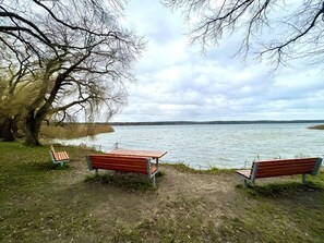 Un área con bancos junto al lago, dos bancos mirando al agua, rodeada de árboles y césped.