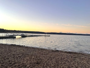 Una playa con un muelle de madera que se adentra en aguas tranquilas, rodeada por una costa arbolada.