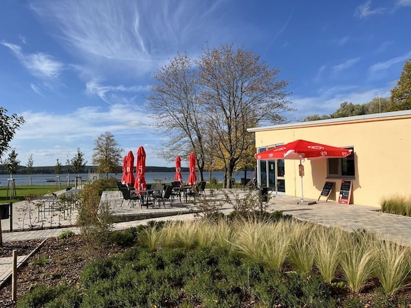 An outdoor seating area with red umbrellas, a building with a yellow facade, and a view of a lake.