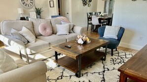 A living room with a sofa, armchair, coffee table, and a dining area in the background.