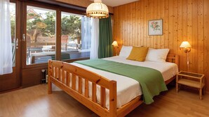 A wooden-framed bed with a green blanket, two bedside lamps, a small wooden chair, and a window with a view of trees. - Centaure B RI 1 by Interhome (Ovronnaz)