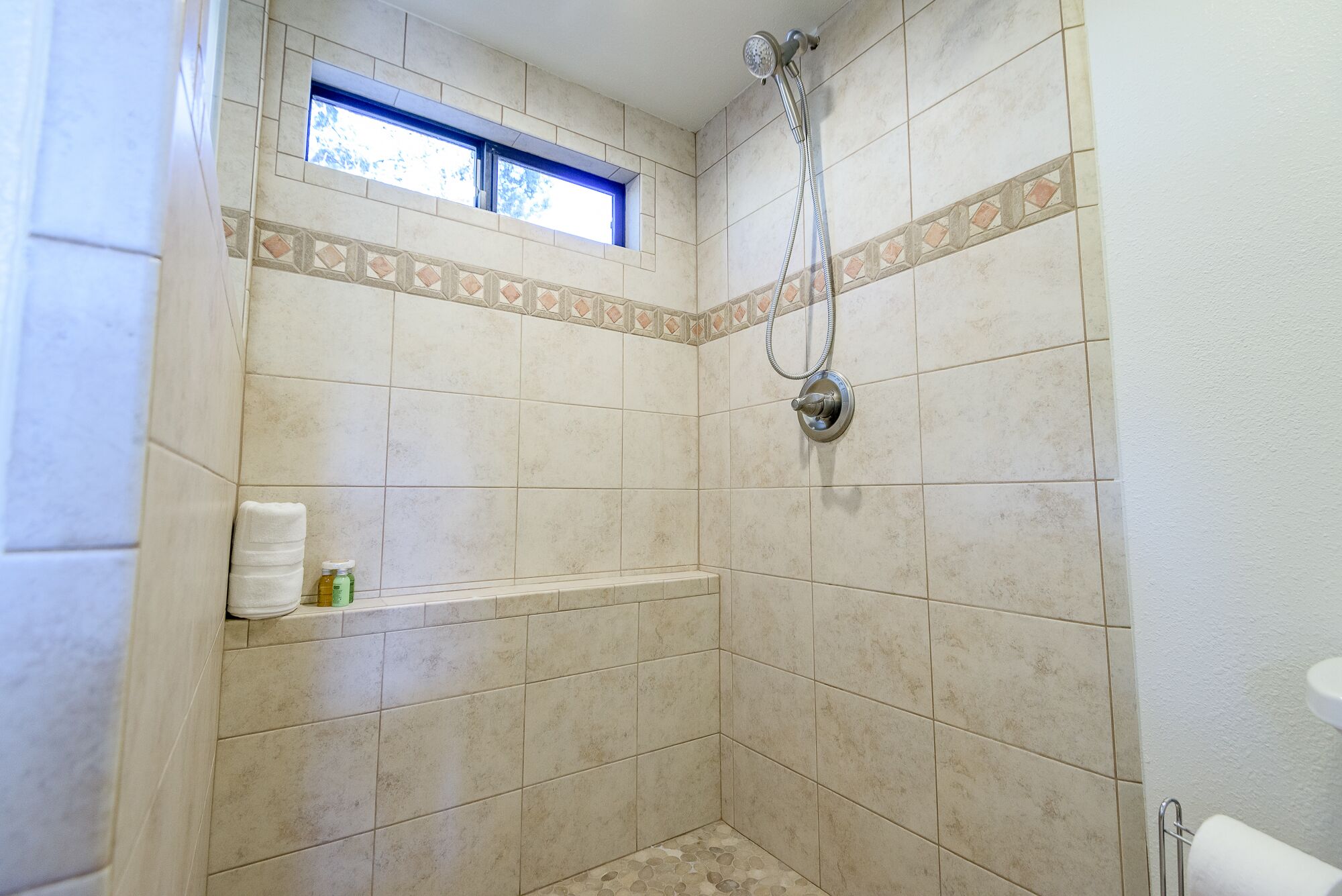 Un espacio de ducha pequeño, con azulejos y una ventana encima, además de un estante con artículos de aseo.
