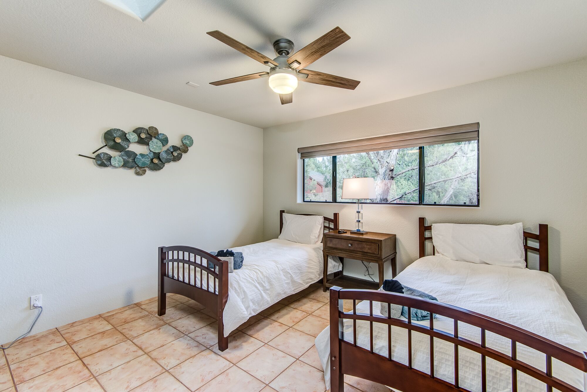 A bedroom with two wooden beds, a ceiling fan, a window with a view, and a wall-mounted lamp.