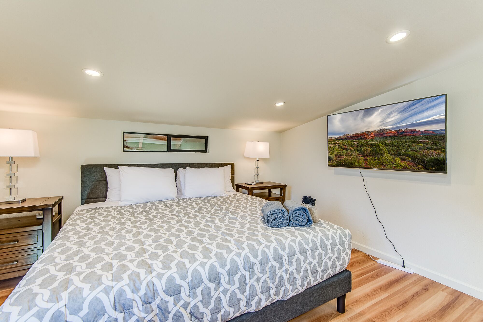 A hotel room with a bed, two bedside tables, a wall-mounted TV, and a painting of a landscape.