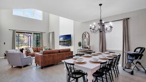 Un amplio salón con una mesa de comedor preparada para seis personas, un sofá marrón, un sillón gris y una araña.