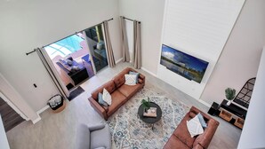 Amplia sala de estar con un sofá marrón, un sillón gris y una mesa de centro. Se aprecia una ventana grande con cortinas, un cuadro en la pared y vistas al exterior.