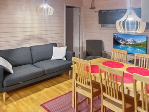 A modern living room with a grey sofa, wooden dining table with red placemats, and a large TV displaying a mountain landscape.