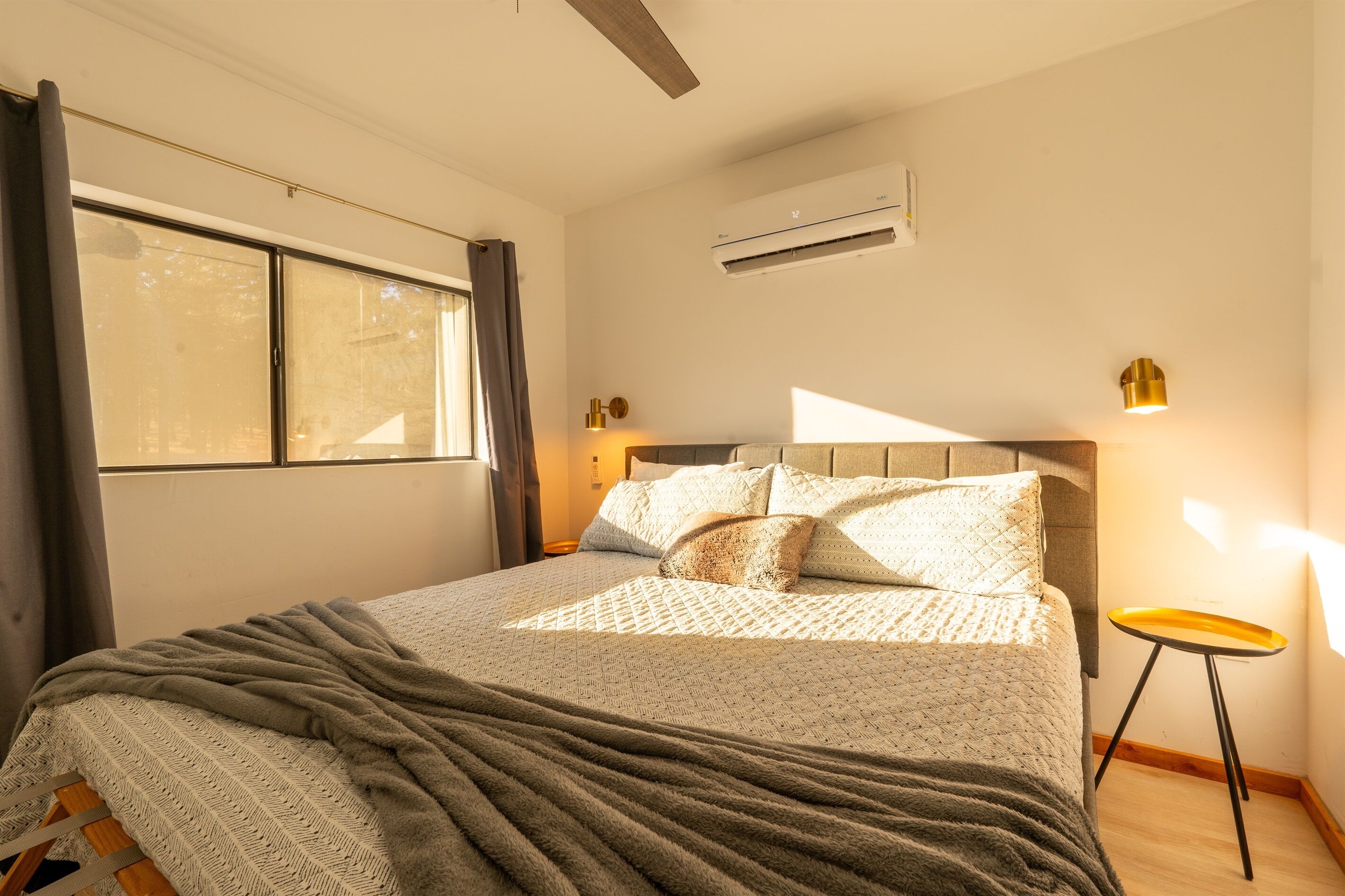 A neatly made bed with a grey blanket and a patterned duvet, a bedside table with a lamp, and a window with curtains.