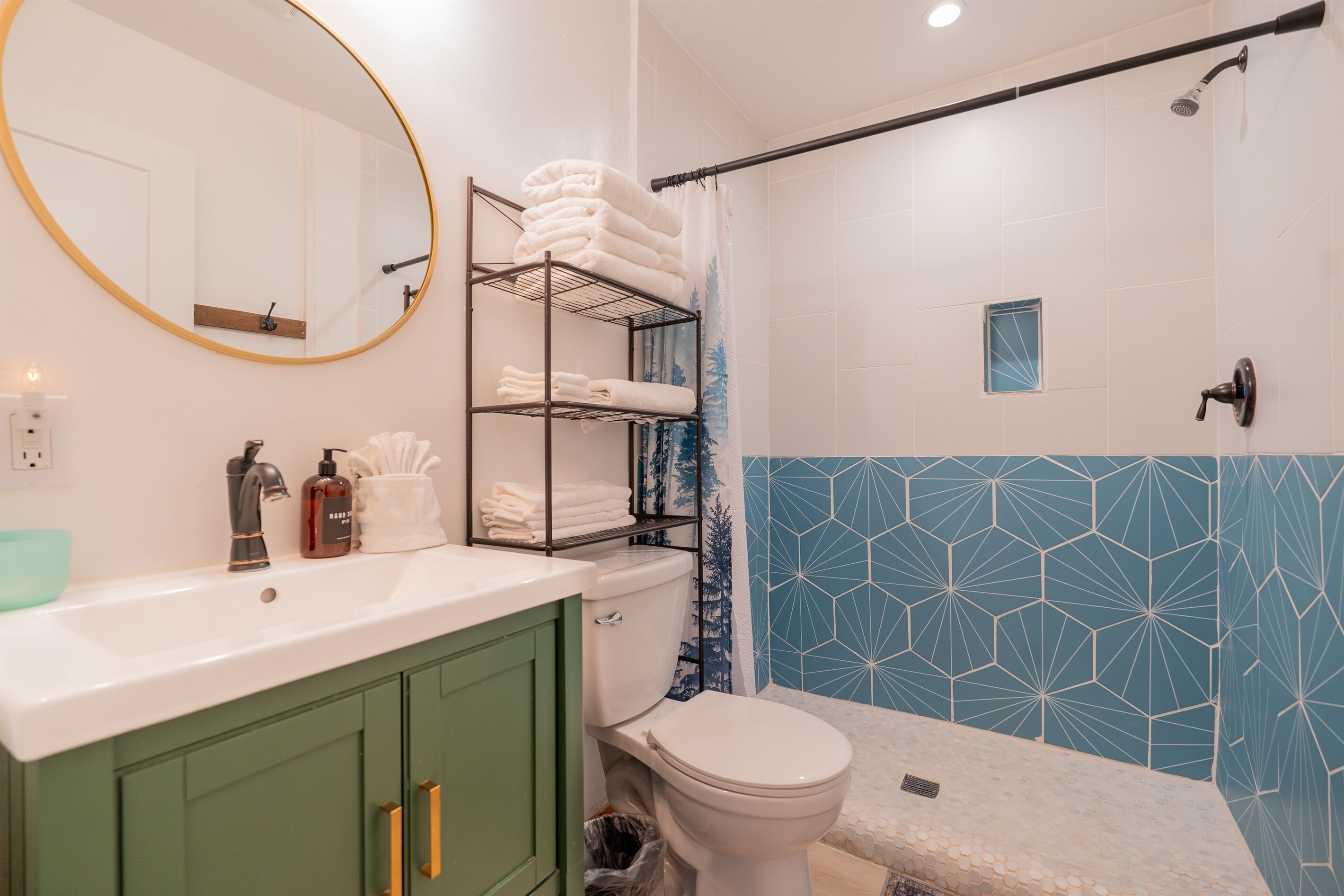 A bathroom with a white toilet, green cabinets, a white sink, a towel rack with folded towels, a shower curtain, and a geometric tile backsplash.