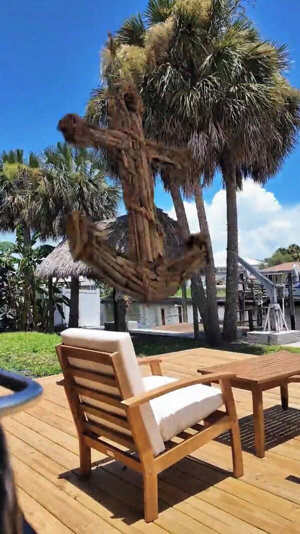 A wooden deck with a lounge chair and a table, surrounded by palm trees and a thatched-roof structure.