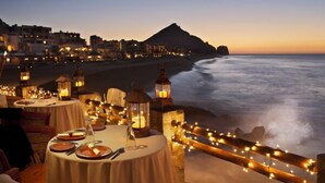 Un restaurant en bord de mer, avec des tables dressées pour le dîner, offrant une vue sur l’océan et un paysage montagneux en arrière-plan.