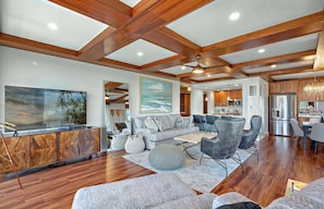 A spacious living room with a large TV, wooden ceiling beams, and a view of the kitchen.