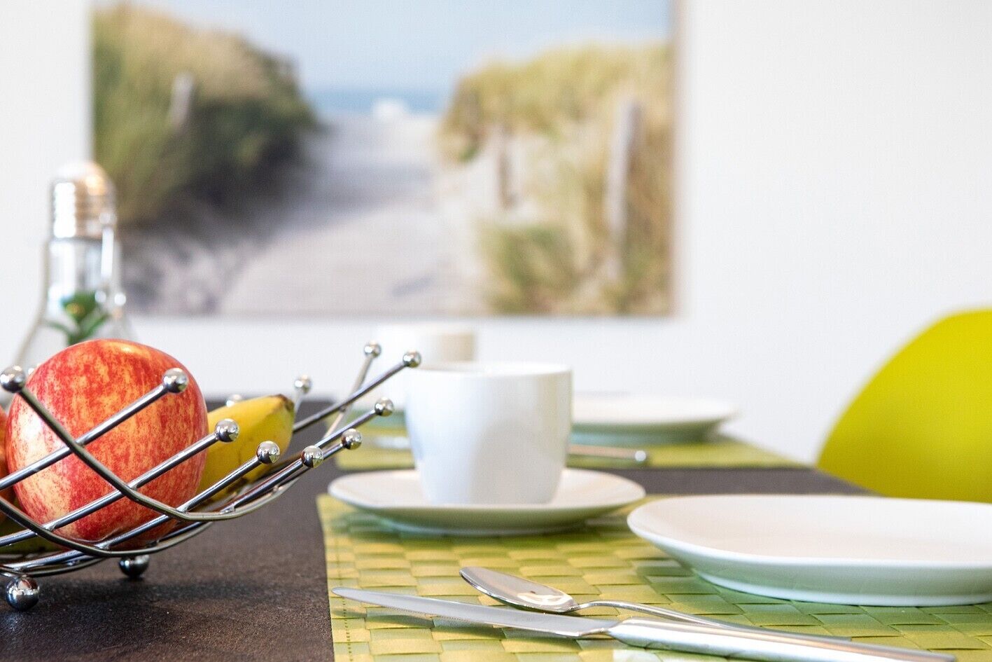 A breakfast setting with a fruit basket, a white cup, and a plate on a table with a woven placemat.