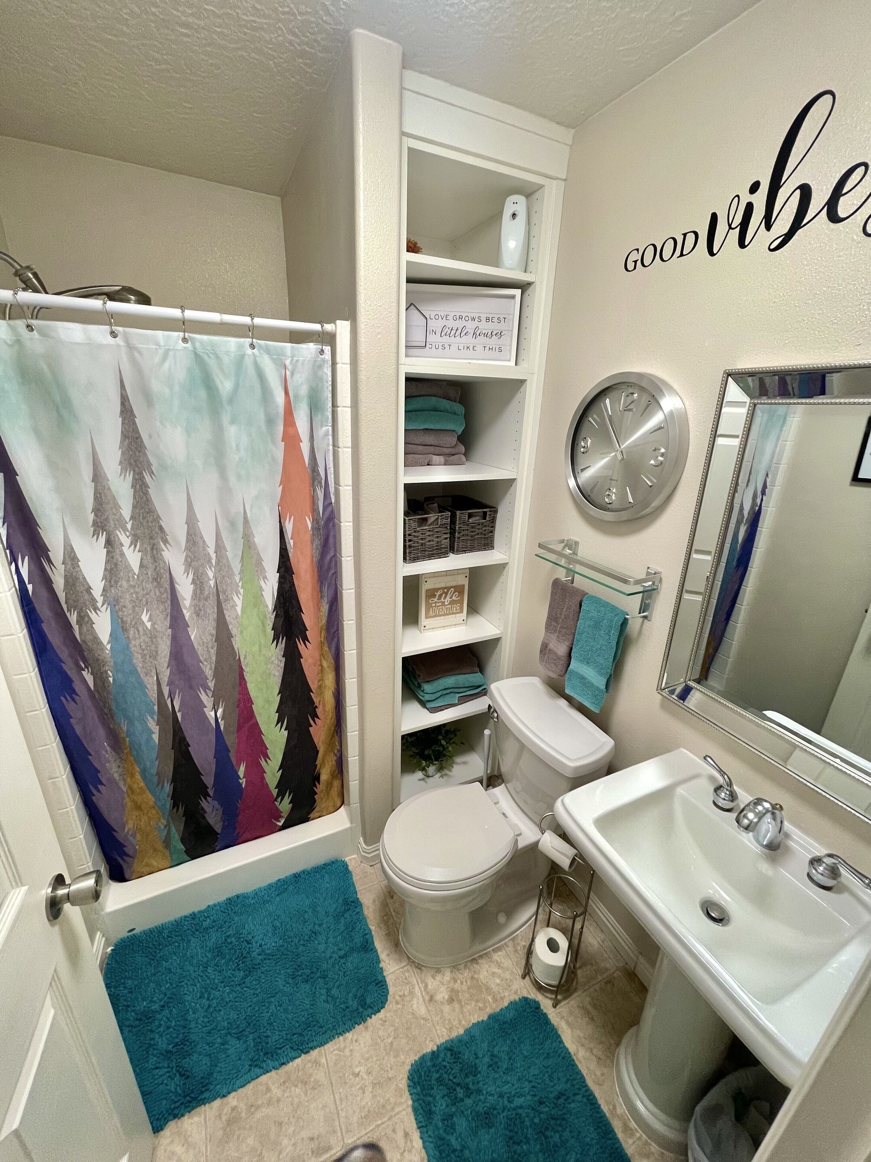 A bathroom with a shower curtain, a toilet, a sink, and a mirror. There are towels and a sign on the shelves.