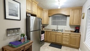 Une cuisine avec des meubles de rangement en bois, un réfrigérateur en acier inoxydable, un évier et une machine à café. On y trouve une petite table en bois sur laquelle sont posés un plateau et des bouteilles.