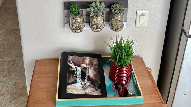 Un cadre contenant le mot « wanderlust » accompagné de sa définition, trois pots suspendus avec des plantes, et une table en bois sur laquelle sont posés un cadre avec une photo et un pot rouge.