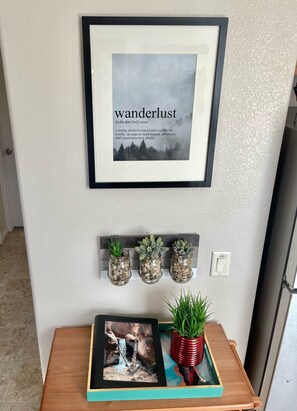 A framed word definition of 'wanderlust' on a wall, a shelf with three potted plants, and a wooden table with a framed photo and a red pot. - Wanderlust House - Cute & Cozy w/ Big Game Room (La Verkin)