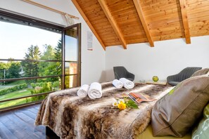 A bedroom with a large window, a bed with rolled towels, a small table with a vase, and a chair.