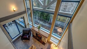 A high-ceilinged room with a fireplace, a seating area, and large windows offering a view of a snowy landscape. - Greystone Lodge 318 Ski-in/Out by Acer Vacations (Whistler)