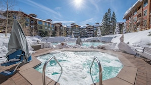 A pool area with a hot tub surrounded by snow, with buildings in the background.