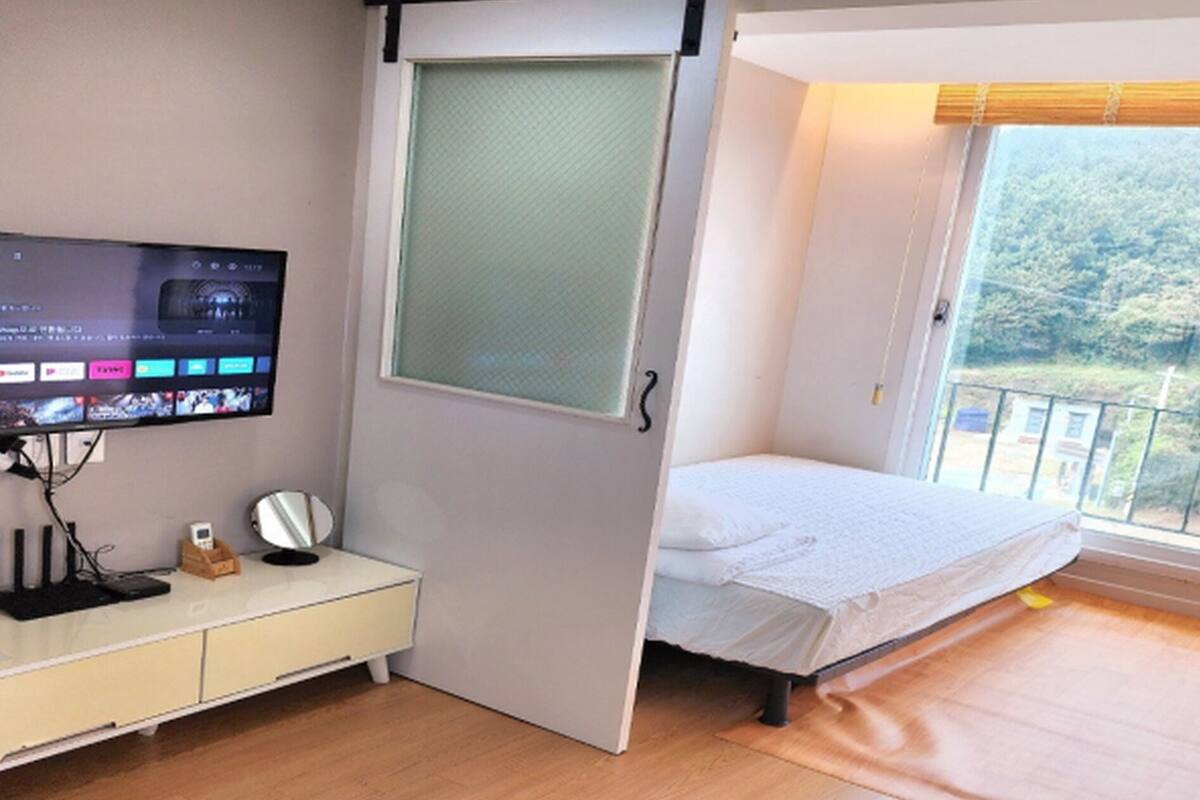 Een slaapkamer met een bed, een televisie, een raam met uitzicht op groen, en een klein bureau met een spiegel.