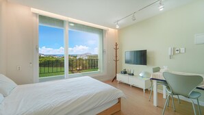 Ein Schlafzimmer mit einem großen Bett, einem Schreibtisch mit Stuhl, einem Fernseher und einem Balkon mit Blick auf Häuser und Grünflächen.