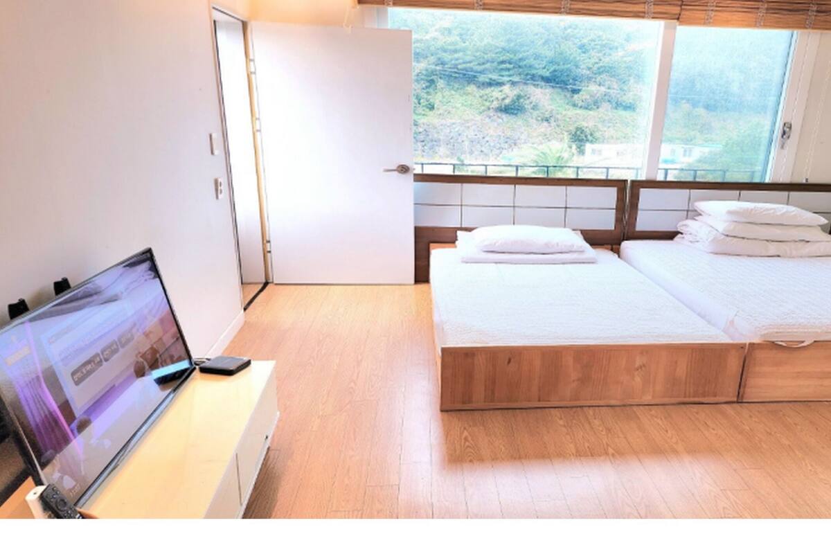 Een slaapkamer met een houten bed, een bureau met een laptop en een raam met uitzicht op groen.