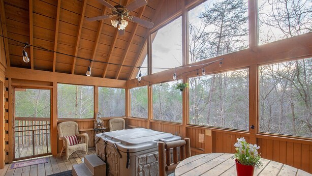 Una cabaña de madera con jacuzzi, sillas de mimbre, una mesa de madera y amplios ventanales con vistas al bosque.