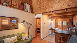 Interior acogedor de una cabaña con un sofá verde, una zona de comedor de madera y una cocina con armarios de madera.