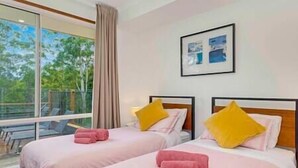A hotel room with two beds, each with pink bedding and a yellow pillow. There is a window with a view of greenery outside.