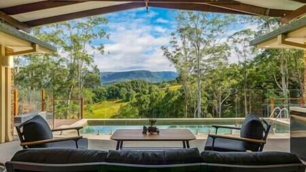 A covered outdoor seating area with a pool, overlooking a lush landscape with trees and mountains.