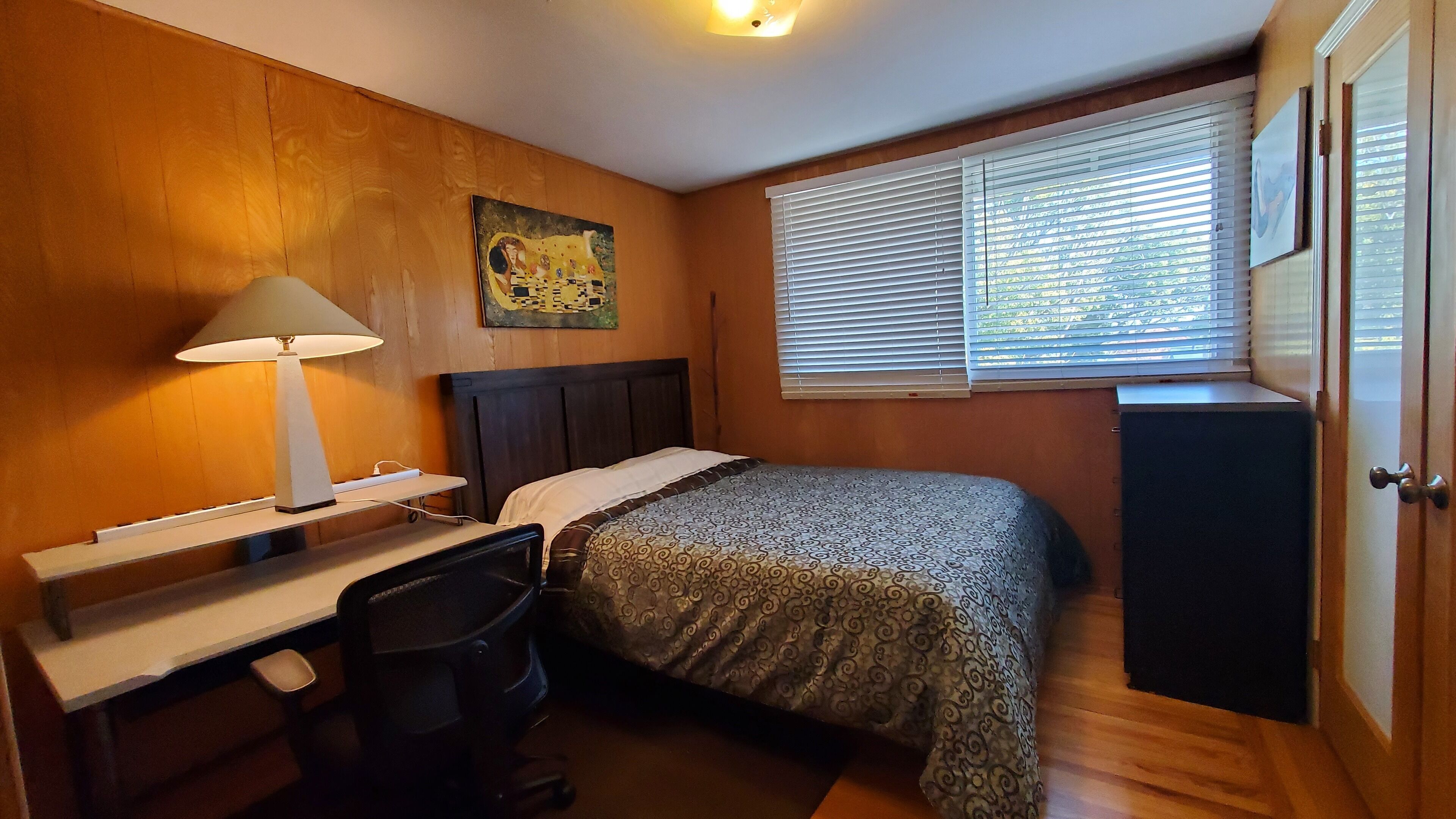 Een slaapkamer met een bed, bureau, stoel, lamp en een schilderij aan de muur.