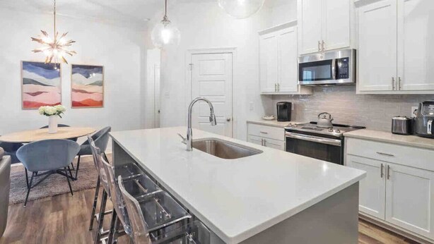 A modern kitchen with white cabinetry, a central island with a sink, and a dining area with a table and chairs. - Cozy & Stylish 3BD near LSU (Baton Rouge)