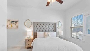 A bedroom with a tufted headboard, a large bed, a nightstand with a lamp, and a window with blinds. - Cozy & Stylish 3BD near LSU (Baton Rouge)