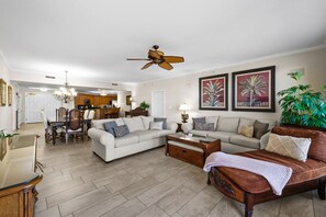 A spacious living room with a large sofa, a wooden coffee table, and a ceiling fan. There is a dining area with a chandelier and a kitchen in the background.