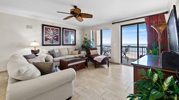 A spacious living room with a sofa, armchairs, a coffee table, and a TV on a wooden stand. There is a ceiling fan, a large window with a view of the ocean, and a balcony with a chair and table.